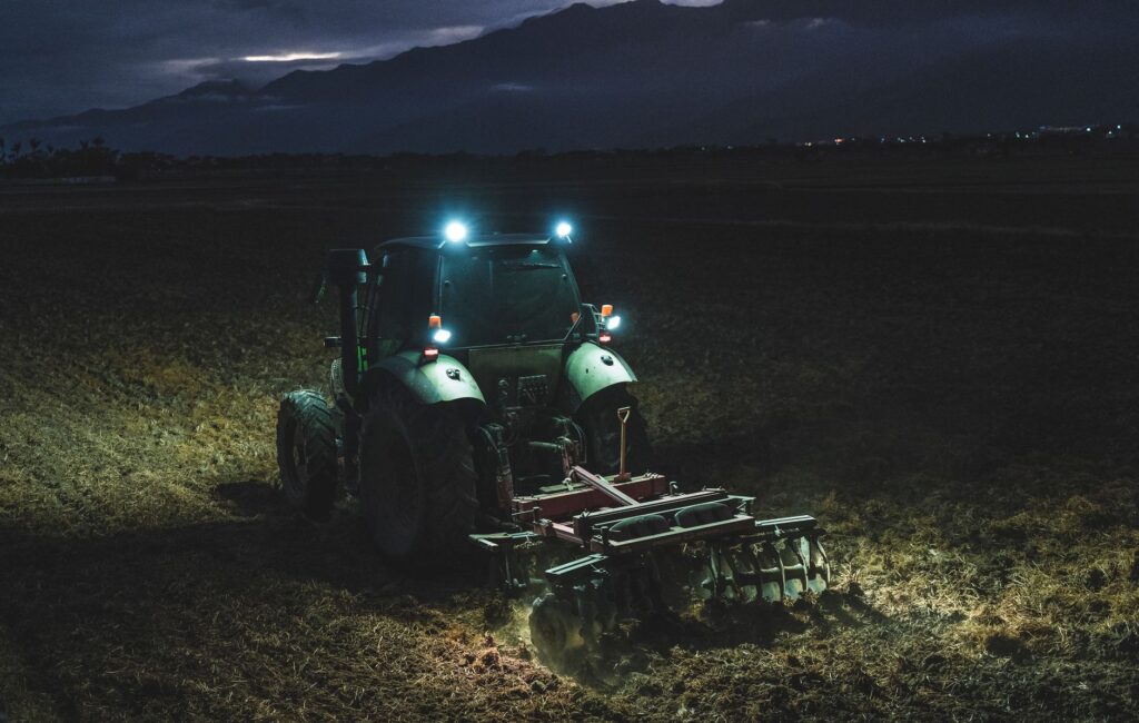 A tractor works in a rural field at night, illuminated by headlights under a cloudy sky.